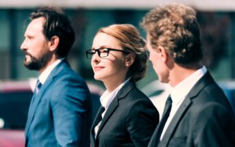 Three professionals, two men and one woman, walk outdoors wearing formal suits. The woman in the center has glasses and a confident expression. They appear engaged in conversation against a blurred urban background.