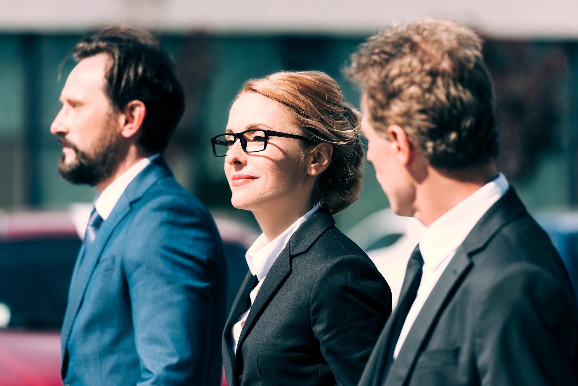 Three professionals, two men and one woman, walk outdoors wearing formal suits. The woman in the center has glasses and a confident expression. They appear engaged in conversation against a blurred urban background.