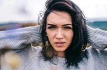 A woman with dark hair and a gray sweater stands outside on a windy day. She holds her hair back with both hands and looks directly at the camera with a focused expression. The background is blurred, suggesting an outdoor setting.