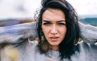 A woman with dark hair and a gray sweater stands outside on a windy day. She holds her hair back with both hands and looks directly at the camera with a focused expression. The background is blurred, suggesting an outdoor setting.