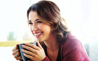 A woman with brown hair smiles while holding a black mug. She is wearing a red sweater and has a ring on her finger. The background is softly lit, suggesting a cozy indoor setting.