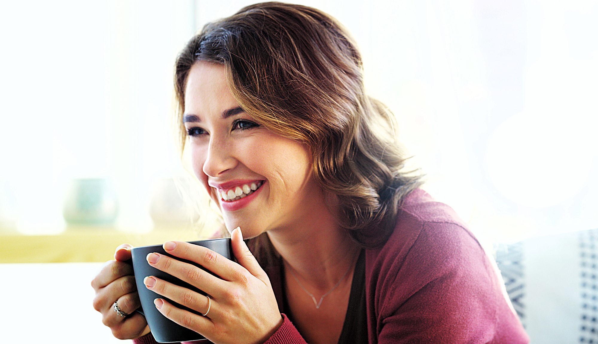 A woman with brown hair smiles while holding a black mug. She is wearing a red sweater and has a ring on her finger. The background is softly lit, suggesting a cozy indoor setting.