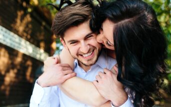 A woman with dark hair playfully hugs a smiling man from behind, wrapping her arms around his shoulders. Both appear to be outside, standing near a brick wall with greenery in the background. The man is wearing a light-colored shirt.