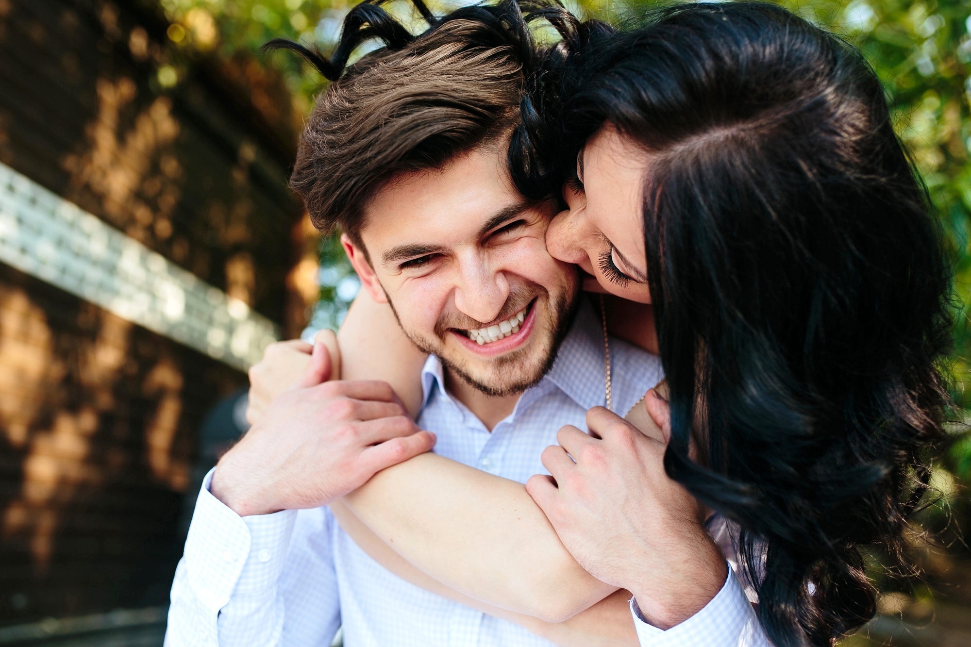 A woman with dark hair playfully hugs a smiling man from behind, wrapping her arms around his shoulders. Both appear to be outside, standing near a brick wall with greenery in the background. The man is wearing a light-colored shirt.