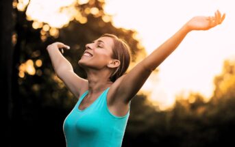 A person with short brown hair is standing outdoors, wearing a blue sleeveless top. They have their arms raised and eyes closed, smiling and facing the warm sunlight. The background is a blurry mix of trees and greenery.