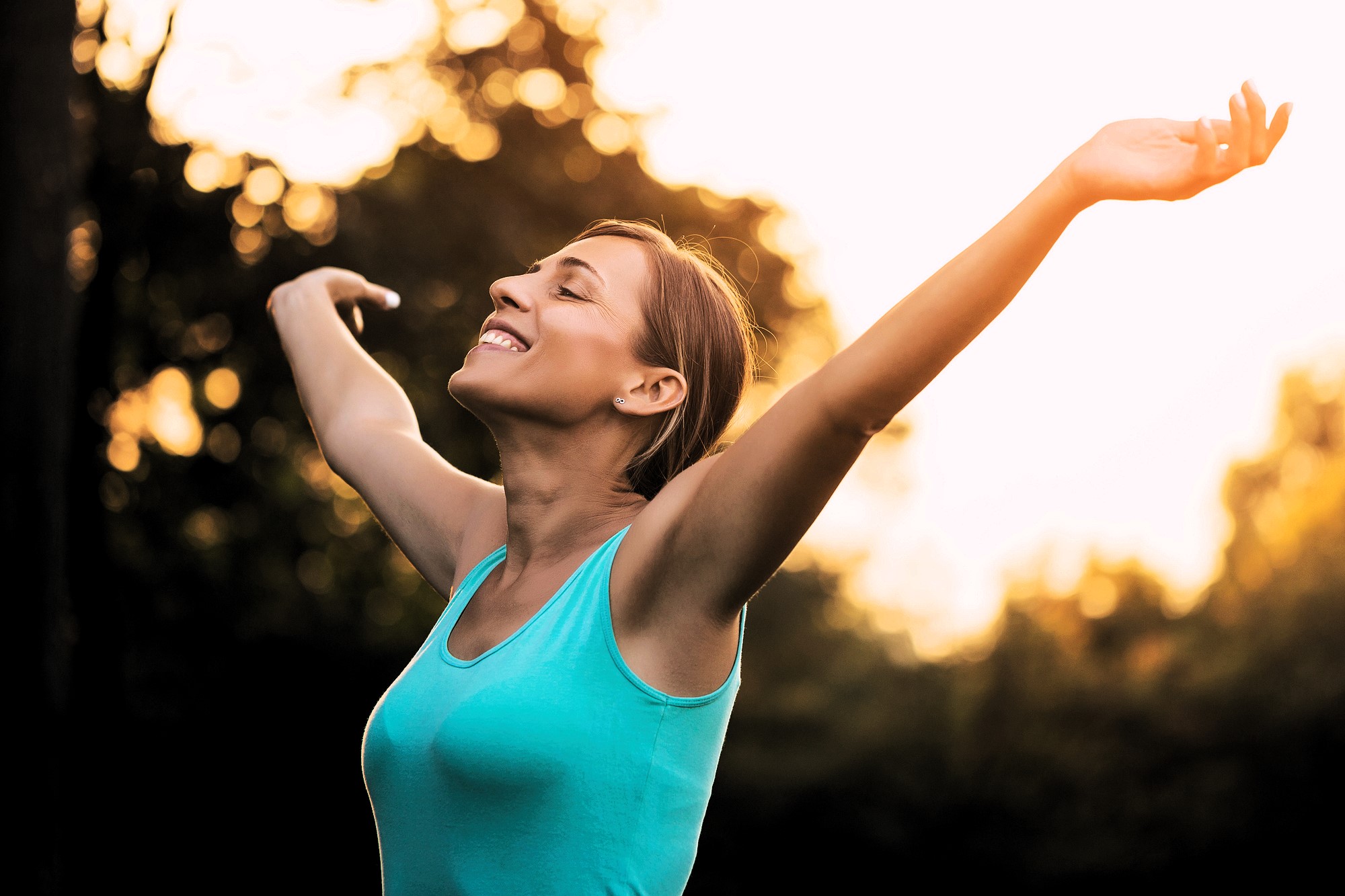 A person with short brown hair is standing outdoors, wearing a blue sleeveless top. They have their arms raised and eyes closed, smiling and facing the warm sunlight. The background is a blurry mix of trees and greenery.