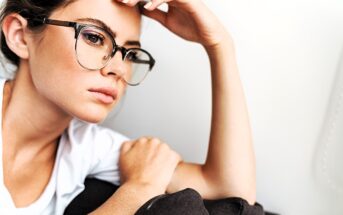 A contemplative individual with glasses and a white shirt rests their chin on a black cushion, gazing off to the side against a light background.