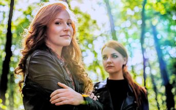 Two women with long red hair stand in a forest with sunlight filtering through the trees. The woman in the foreground wears a leather jacket and looks towards the camera, while the other gazes to the side.