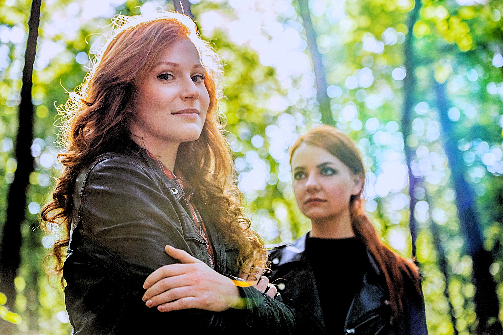 Two women with long red hair stand in a forest with sunlight filtering through the trees. The woman in the foreground wears a leather jacket and looks towards the camera, while the other gazes to the side.