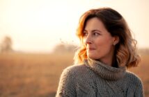 A woman with shoulder-length hair stands in a field wearing a thick knit sweater. She is gazing to her left with a small, thoughtful smile. The sun is setting, casting a warm glow over the scene.