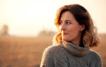 A woman with shoulder-length hair stands in a field wearing a thick knit sweater. She is gazing to her left with a small, thoughtful smile. The sun is setting, casting a warm glow over the scene.