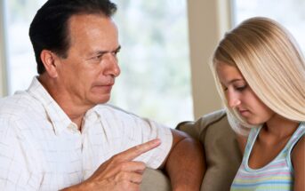 A man in a white shirt is sitting on a couch, pointing his finger while speaking to a young woman with long blonde hair wearing a striped tank top. The woman appears to be looking down, showing a serious expression.