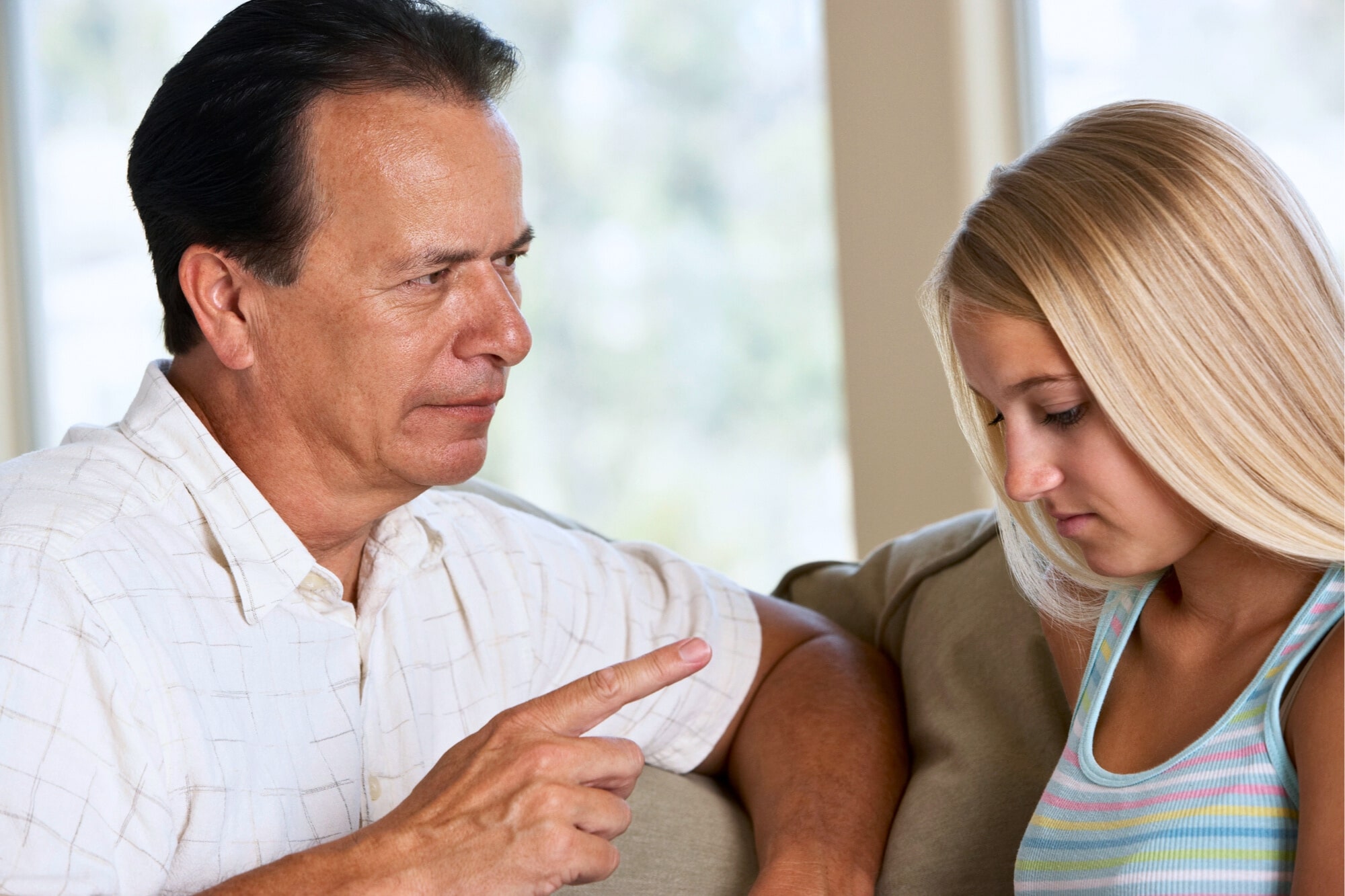 A man in a white shirt is sitting on a couch, pointing his finger while speaking to a young woman with long blonde hair wearing a striped tank top. The woman appears to be looking down, showing a serious expression.