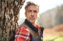 A man with gray hair and a beard, wearing a red plaid shirt and a vest, leans against a tree outdoors. He looks thoughtfully into the distance with a blurred natural background behind him.