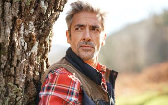 A man with gray hair and a beard, wearing a red plaid shirt and a vest, leans against a tree outdoors. He looks thoughtfully into the distance with a blurred natural background behind him.