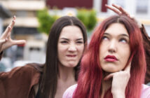 A woman with red hair looks upward and holds her chin in thought, while another woman with dark hair in the background makes an exaggerated expression, appearing to imitate claws with her hands. They stand outdoors, and the background is slightly blurred.