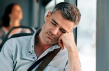 A man with gray hair is sitting on a bus, resting his head on his hand against the window, appearing to be sleeping. He is wearing a light button-up shirt and has a strap across his shoulder. The background shows an unfocused passenger.