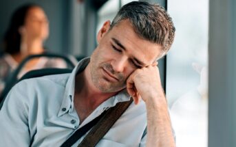 A man with gray hair is sitting on a bus, resting his head on his hand against the window, appearing to be sleeping. He is wearing a light button-up shirt and has a strap across his shoulder. The background shows an unfocused passenger.