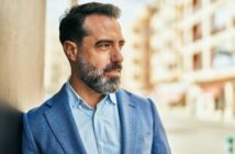 A man with a beard and salt-and-pepper hair stands outdoors, leaning against a wall. He is wearing a blue blazer over a light blue shirt and is looking to the side, with buildings blurred in the background.