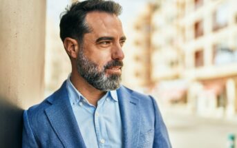 A man with a beard and salt-and-pepper hair stands outdoors, leaning against a wall. He is wearing a blue blazer over a light blue shirt and is looking to the side, with buildings blurred in the background.