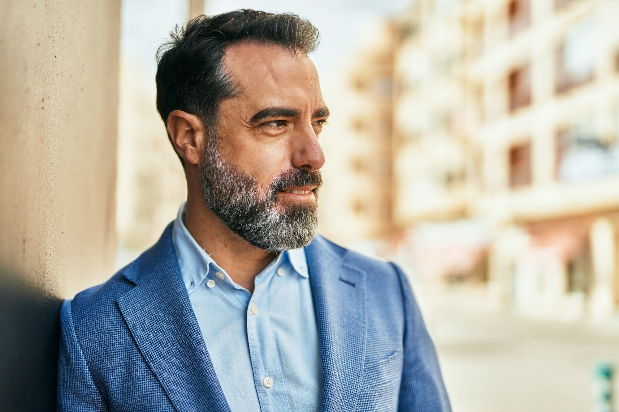 A man with a beard and salt-and-pepper hair stands outdoors, leaning against a wall. He is wearing a blue blazer over a light blue shirt and is looking to the side, with buildings blurred in the background.