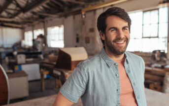 A man in a light denim shirt smiles while standing in a well-lit workshop. The background features tools, equipment, and large windows, creating a spacious and industrious atmosphere.