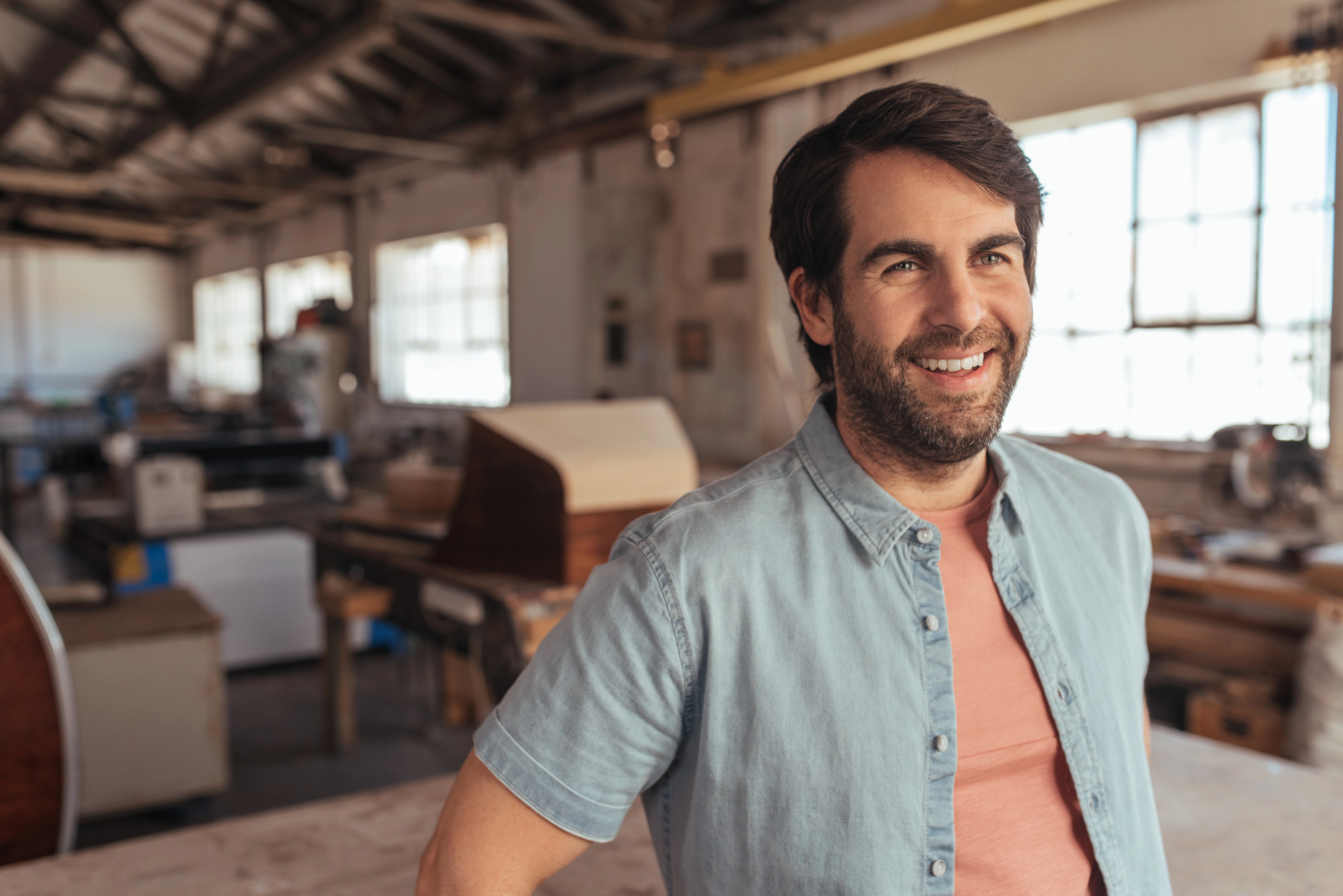 A man in a light denim shirt smiles while standing in a well-lit workshop. The background features tools, equipment, and large windows, creating a spacious and industrious atmosphere.