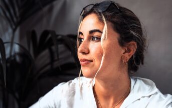 A woman with wavy hair and a white shirt looks to the side with a thoughtful expression. She wears earrings and sunglasses on her head. The background features dark green leaves and a gray wall.