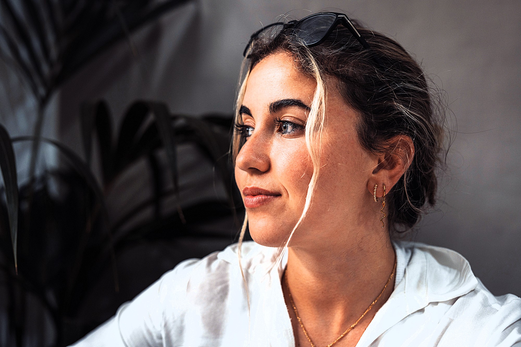 A woman with wavy hair and a white shirt looks to the side with a thoughtful expression. She wears earrings and sunglasses on her head. The background features dark green leaves and a gray wall.