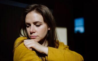 A woman with long brown hair looks down thoughtfully, resting her head on her folded arm. She is wearing a mustard yellow top, and the sunlight creates dramatic shadows on her face and the background.