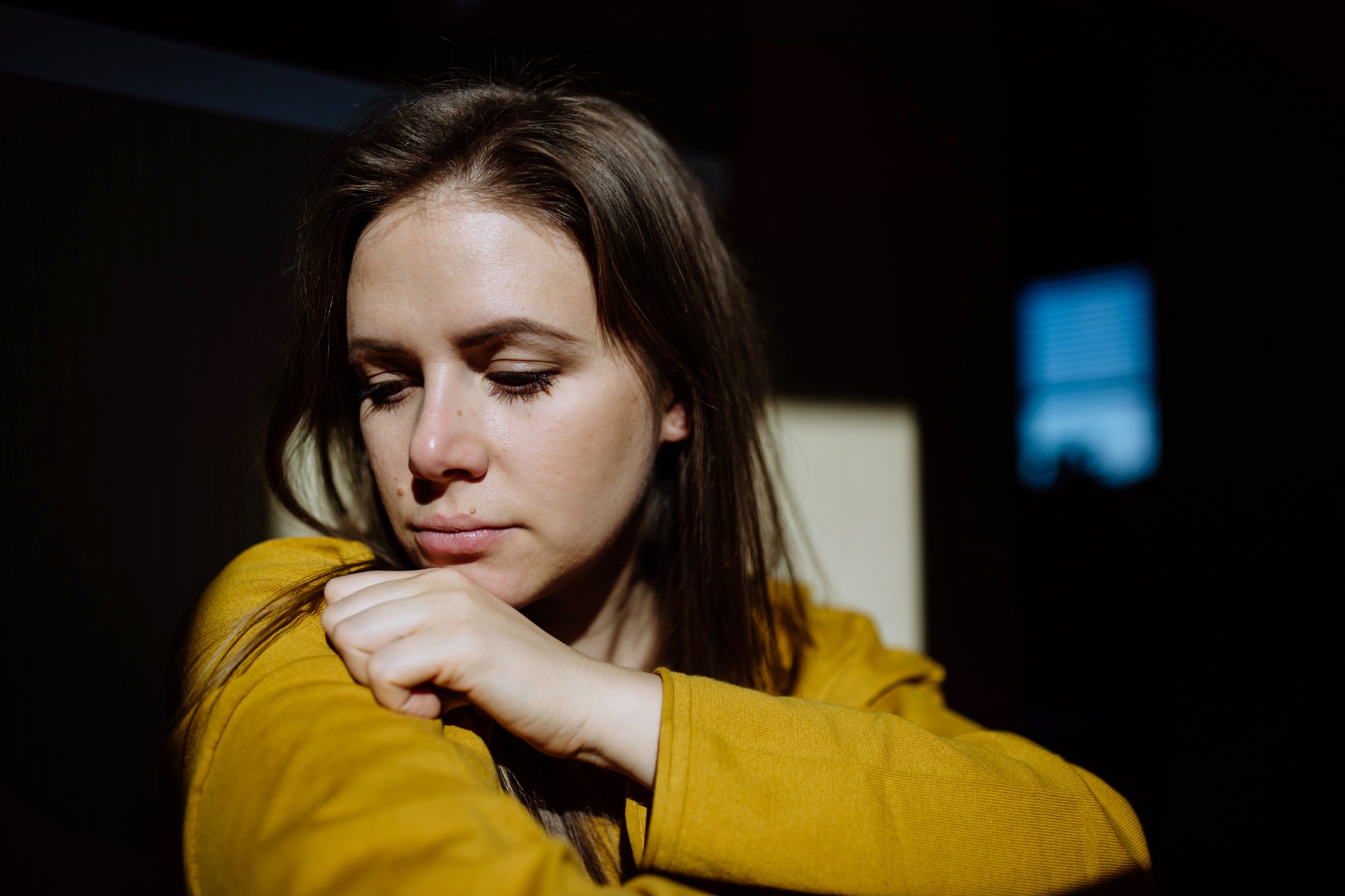 A woman with long brown hair looks down thoughtfully, resting her head on her folded arm. She is wearing a mustard yellow top, and the sunlight creates dramatic shadows on her face and the background.