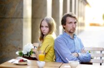 A woman and a man sitting back-to-back at an outdoor café table, arms crossed. She wears a yellow jacket, and he wears a blue shirt. The table has drinks and dishes, and the background features a sunlit corridor.