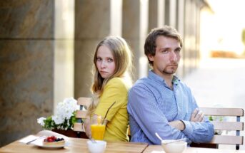 A woman and a man sitting back-to-back at an outdoor café table, arms crossed. She wears a yellow jacket, and he wears a blue shirt. The table has drinks and dishes, and the background features a sunlit corridor.