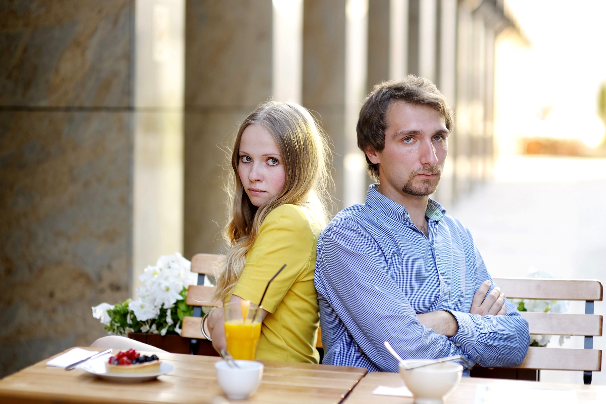 A woman and a man sitting back-to-back at an outdoor café table, arms crossed. She wears a yellow jacket, and he wears a blue shirt. The table has drinks and dishes, and the background features a sunlit corridor.