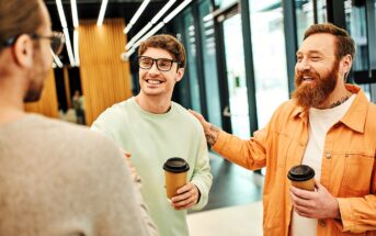 Three men are standing indoors, smiling and talking. Two of them are holding coffee cups. The man on the right, wearing an orange jacket and with tattoos, is patting the shoulder of another man in glasses and a light green sweater.