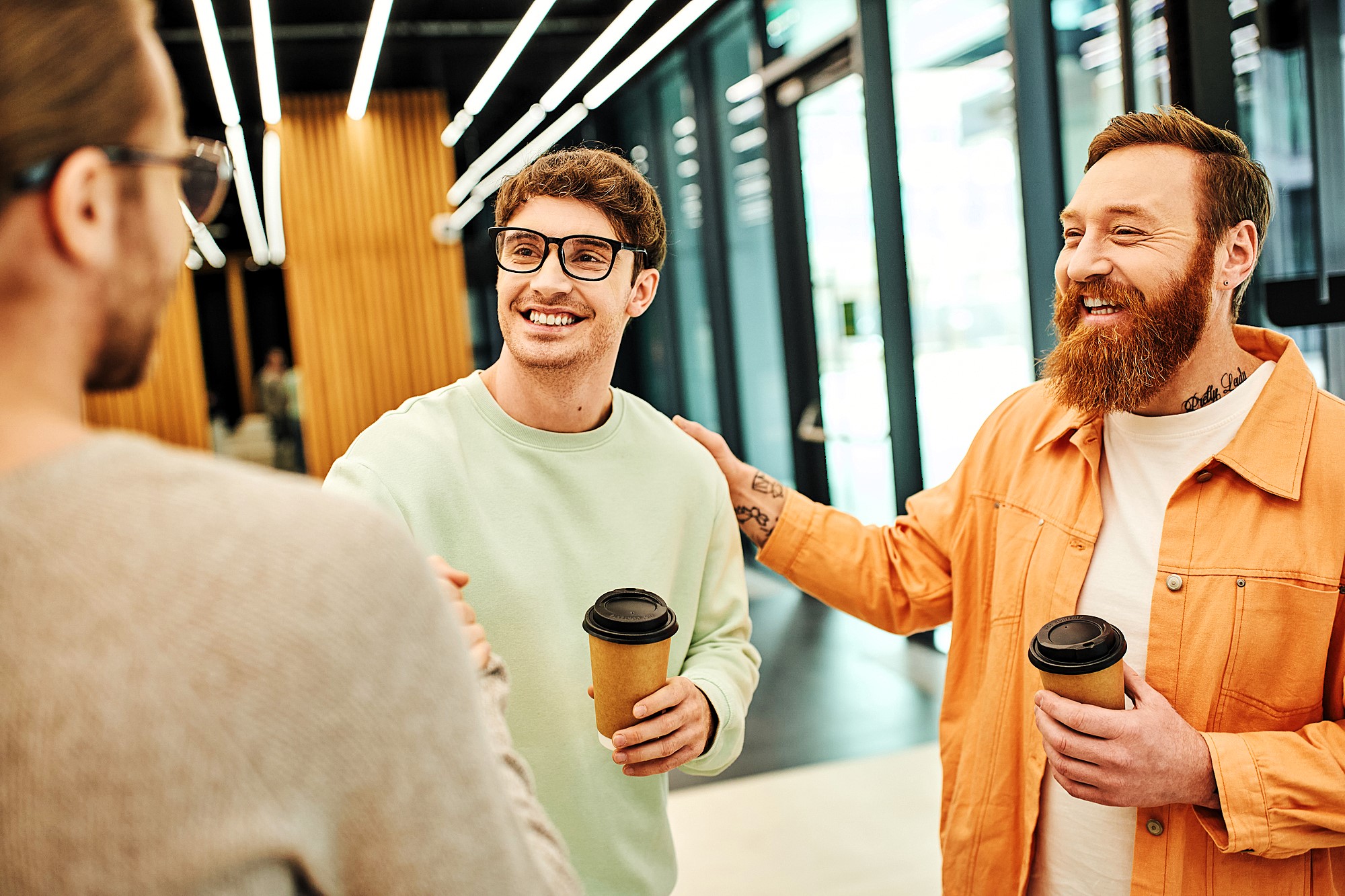 Three men are standing indoors, smiling and talking. Two of them are holding coffee cups. The man on the right, wearing an orange jacket and with tattoos, is patting the shoulder of another man in glasses and a light green sweater.