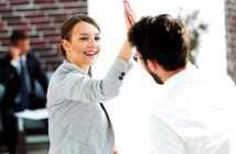 A woman in a gray blazer and a man in a white shirt share a high-five, smiling in a bright office setting. A person in the background appears to be talking on a phone.