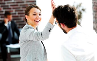 A woman in a gray blazer and a man in a white shirt share a high-five, smiling in a bright office setting. A person in the background appears to be talking on a phone.