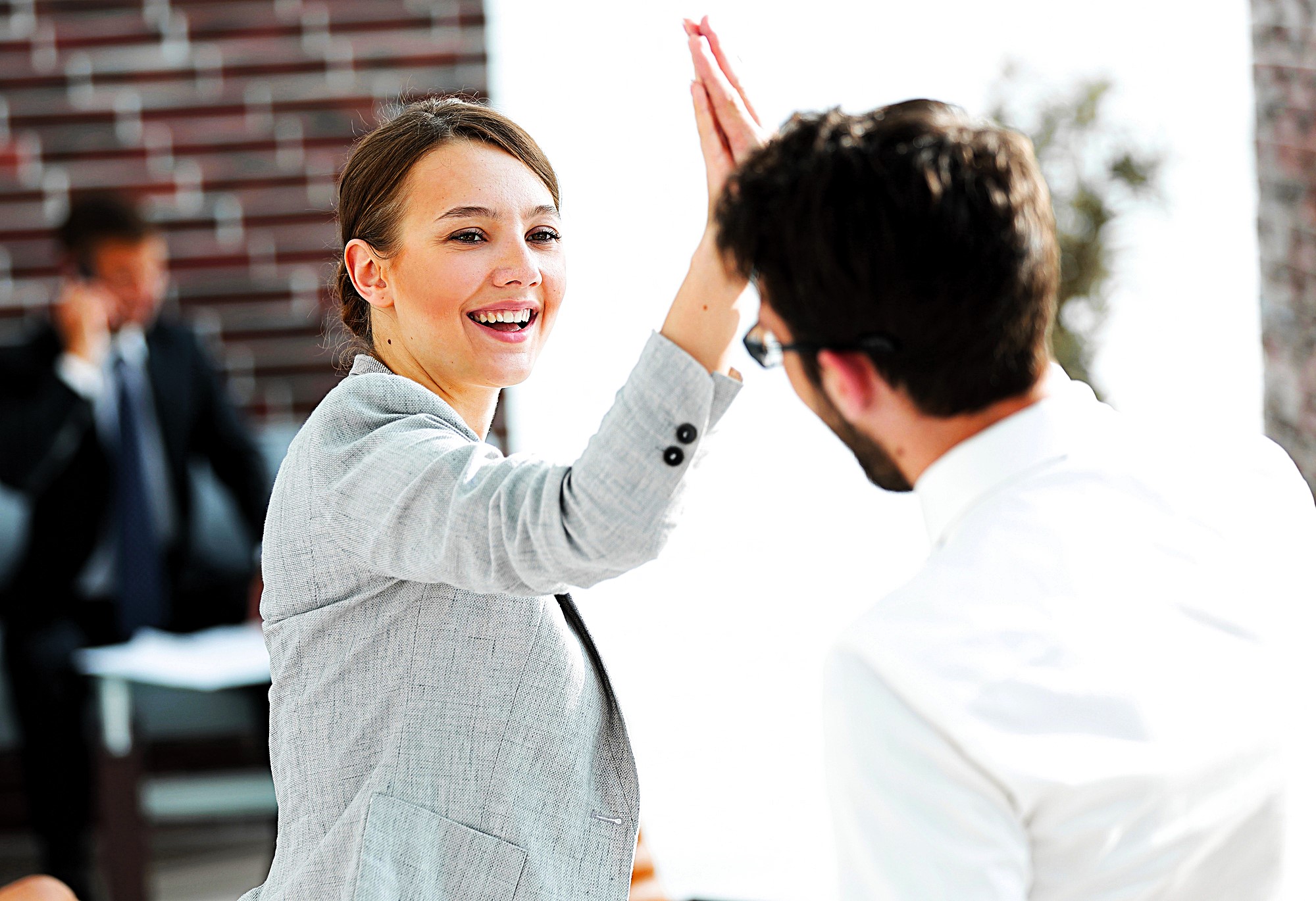 A woman in a gray blazer and a man in a white shirt share a high-five, smiling in a bright office setting. A person in the background appears to be talking on a phone.