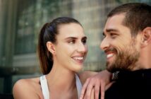 A woman and a man smiling at each other with affection. She has long hair tied back and is wearing a white sleeveless top, while he has a beard and is wearing a black jacket. They are outdoors, with a building in the background.