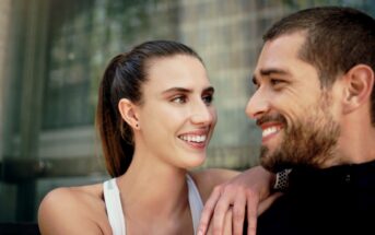 A woman and a man smiling at each other with affection. She has long hair tied back and is wearing a white sleeveless top, while he has a beard and is wearing a black jacket. They are outdoors, with a building in the background.