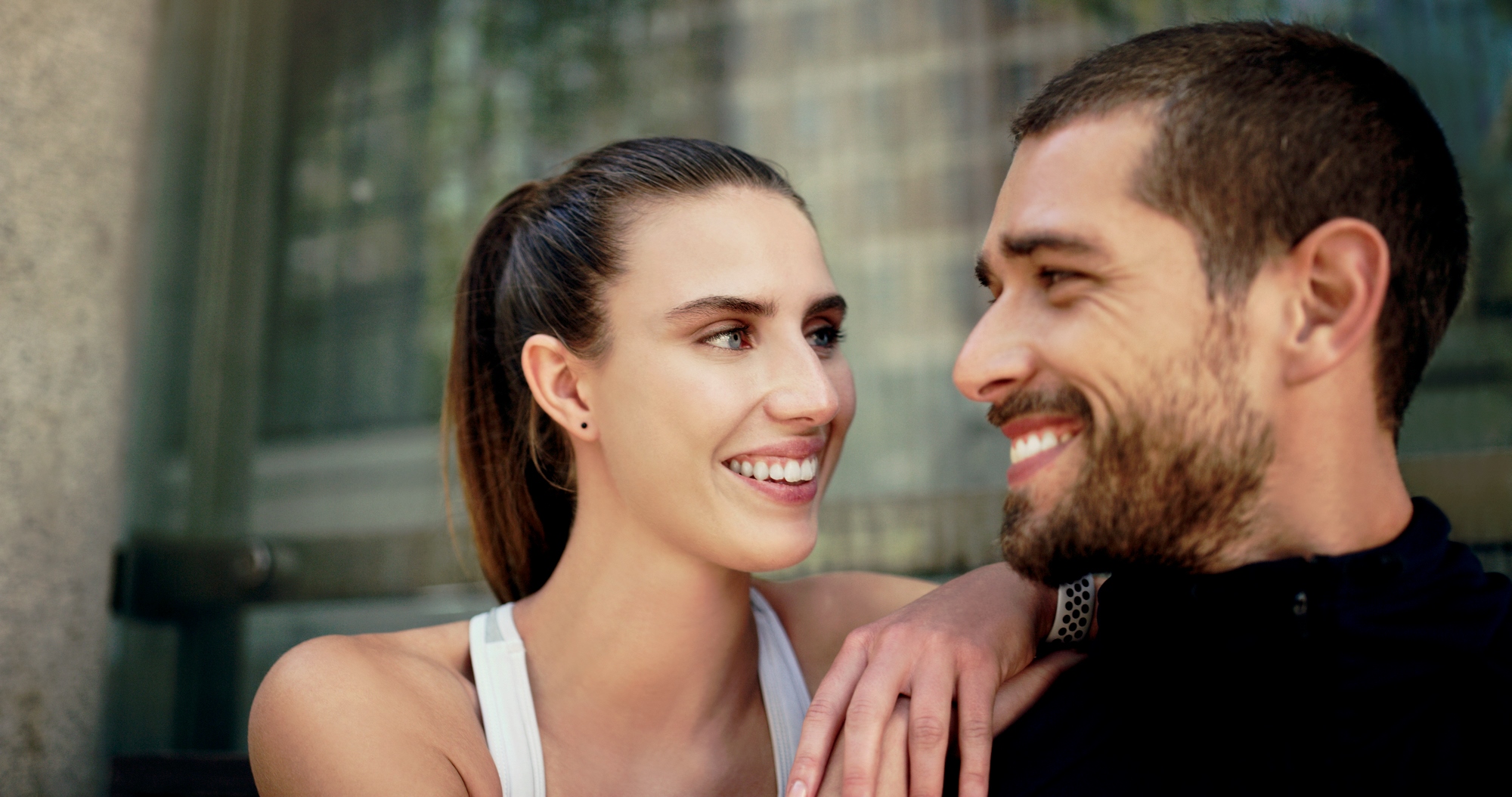 A woman and a man smiling at each other with affection. She has long hair tied back and is wearing a white sleeveless top, while he has a beard and is wearing a black jacket. They are outdoors, with a building in the background.