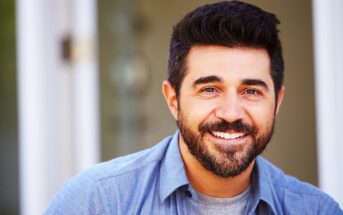 Man with short dark hair and a beard smiling warmly, wearing a light blue collared shirt over a gray t-shirt. The background is softly blurred, suggesting an indoor setting.