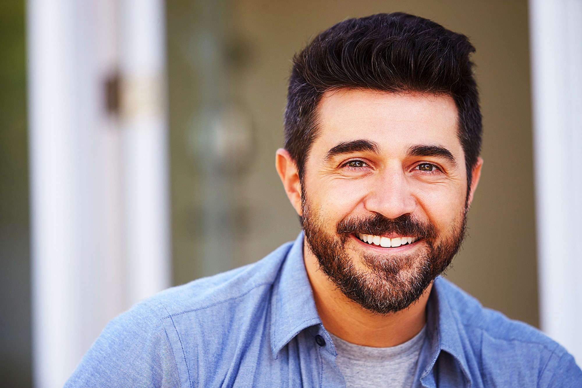 Man with short dark hair and a beard smiling warmly, wearing a light blue collared shirt over a gray t-shirt. The background is softly blurred, suggesting an indoor setting.