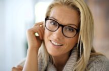 A person with long blonde hair and glasses is smiling warmly. They are wearing a cozy, light-colored sweater and resting their hand on their cheek, set against a softly blurred background.