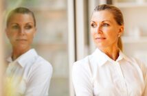 A thoughtful woman in a white blouse stands by a window, gazing outside. Her reflection is visible on the glass next to her. The background is softly blurred, adding a serene atmosphere to the image.