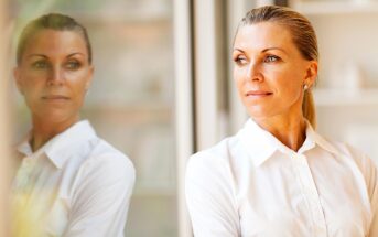 A thoughtful woman in a white blouse stands by a window, gazing outside. Her reflection is visible on the glass next to her. The background is softly blurred, adding a serene atmosphere to the image.