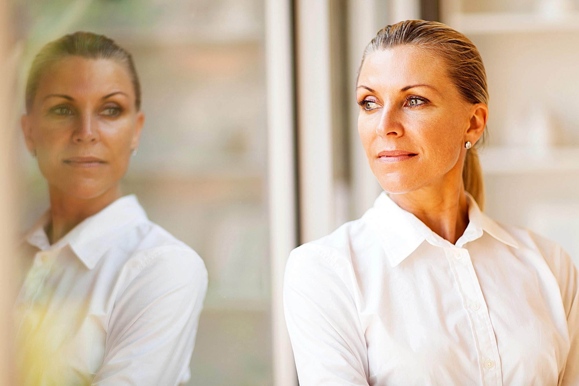 A thoughtful woman in a white blouse stands by a window, gazing outside. Her reflection is visible on the glass next to her. The background is softly blurred, adding a serene atmosphere to the image.