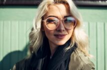 A person with long blonde hair is wearing large, round glasses and a dark scarf, smiling slightly while leaning against a green panel wall in bright sunlight.