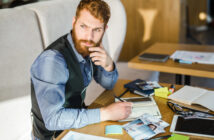 A bearded man in a blue shirt and black vest sits at a table filled with papers, photos, post-it notes, and electronic devices. He looks thoughtful, resting his hand on his chin as he holds a pen over an open notebook.